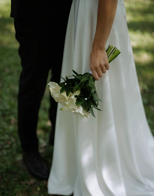 Çimlerle kaplı bir açık hava mekanında, siyah takım elbiseli damadın yanında duran, beyaz kalla zambaklarından oluşan bir buket tutan, dalgalı beyaz bir gelinlik giymiş gelin.