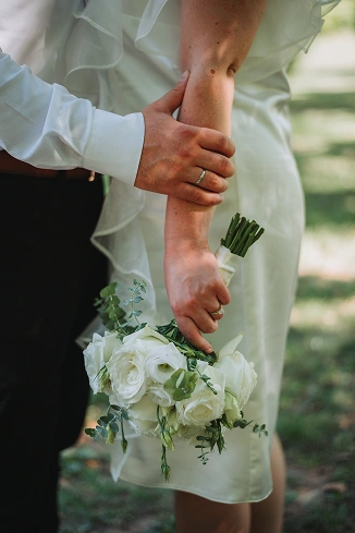 Beyaz güller ve yeşilliklerden oluşan bir buket tutan gelinin yakın çekimi; damadın eli gelinin koluna nazikçe dokunuyor. Sahne, şefkat dolu ve samimi.