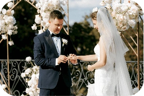 Lacivert smokin giymiş bir damat, açık havada düzenlenen bir düğün sırasında gelinin parmağına yüzüğü takıyor. Beyaz gelinlik ve duvak giymiş gelin, çiçek süslemelerinin arasında gülümsüyor.