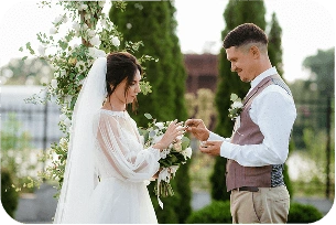 Bir açık hava düğün töreninde gelin ve damat yüzüklerini takas ediyorlar. Çiçeklerden yapılmış bir kemerin altında duruyorlar ve birbirlerine şefkatle gülümsüyorlar.